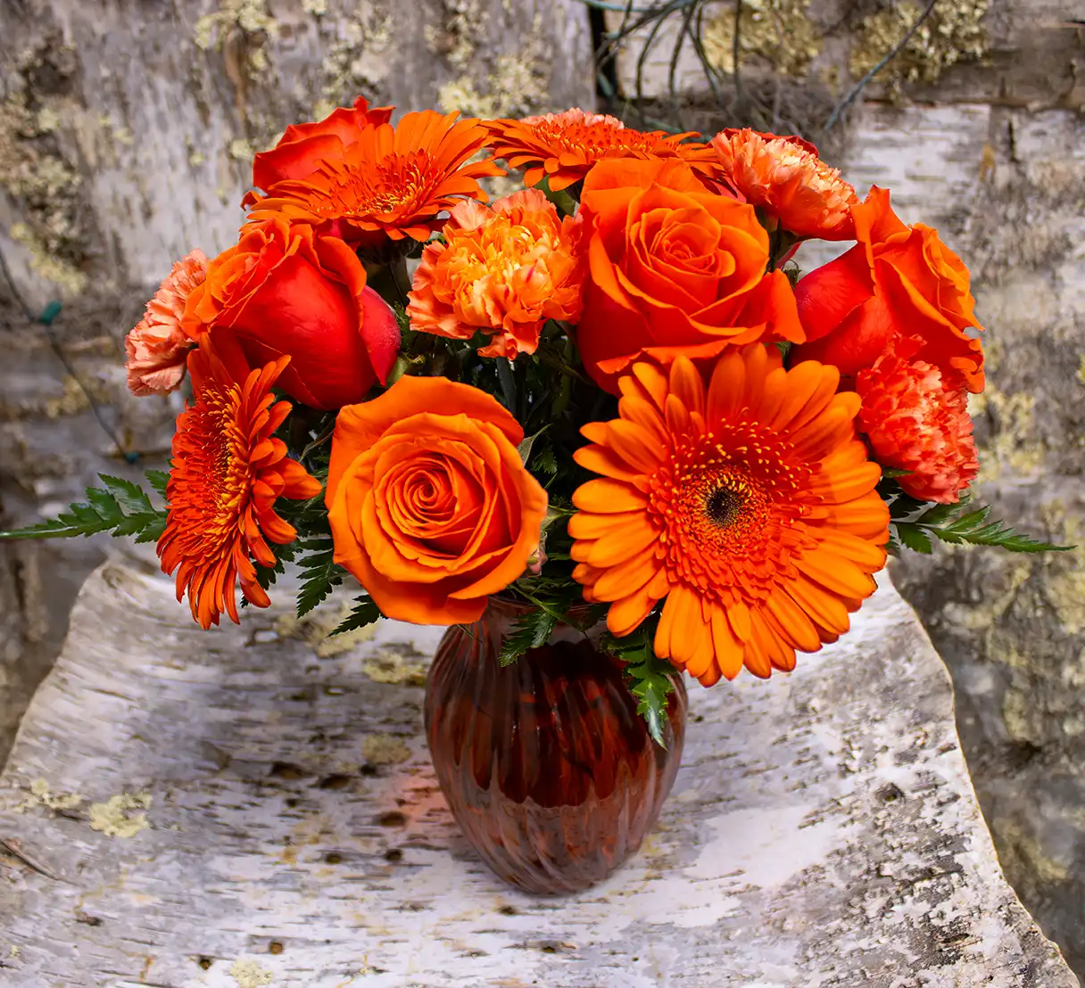 This vibrant floral bouquet features a harmonious collection of all-orange roses, Gerber daisies, and carnations, expertly arranged in a striking orange glass vase. The roses bring a classic elegance, while the Gerber daisies add a cheerful and radiant touch. The carnations complement with their soft, ruffled texture, creating a delightful mix of shapes and forms. The orange vase enhances the monochromatic theme, tying the bouquet together in a warm, bold display perfect for brightening any space.