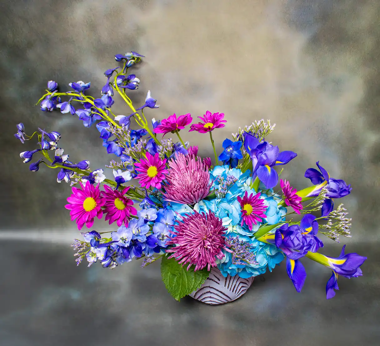 A captivating floral arrangement of purple and blue blossoms exudes charm and elegance. The bouquet features delicate purple daisies interwoven with the striking vertical spikes of blue delphinium, creating a sense of height and grace. Velvety clusters of hydrangea in soft lavender hues provide a lush and textural backdrop, harmonizing beautifully with the dynamic curves of vibrant blue iris blooms.